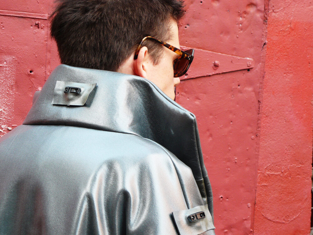 A close-up rear view of a person wearing a metallic silver garment with tag-like fastenings, against a red textured wall.