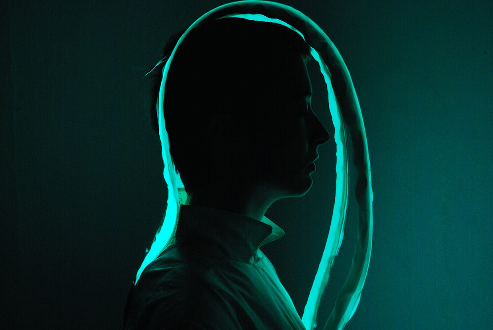 A dark, silhouetted figure in profile with a glowing teal light outlining the head and shoulders, resembling a hood.