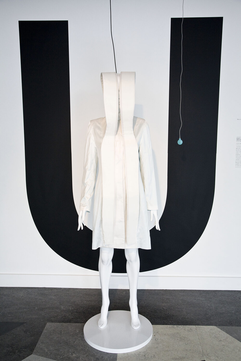 A pale, hooded garment displayed on a mannequin, folded fabric rising above the head, against a black letter U on the wall behind.