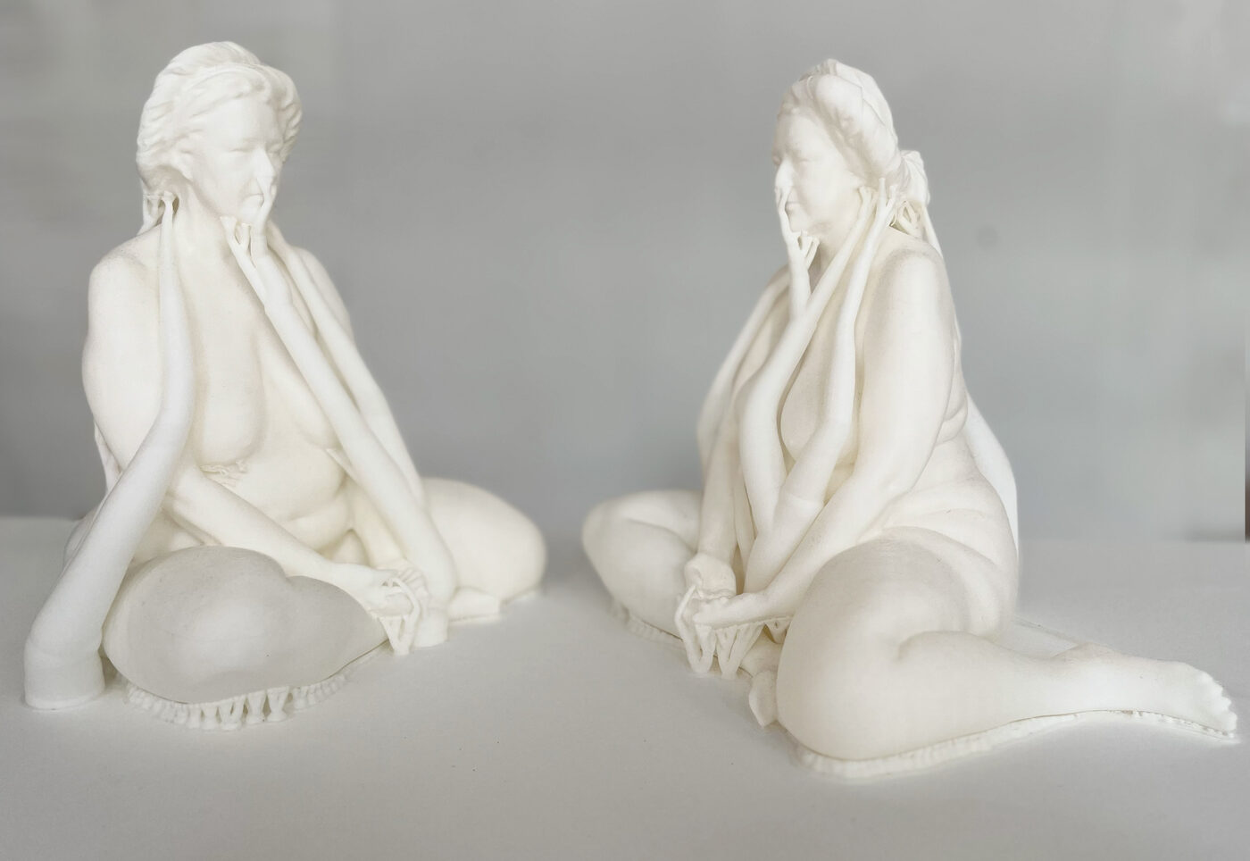 Two pale seated sculptural figures facing one another, hands raised toward their faces.