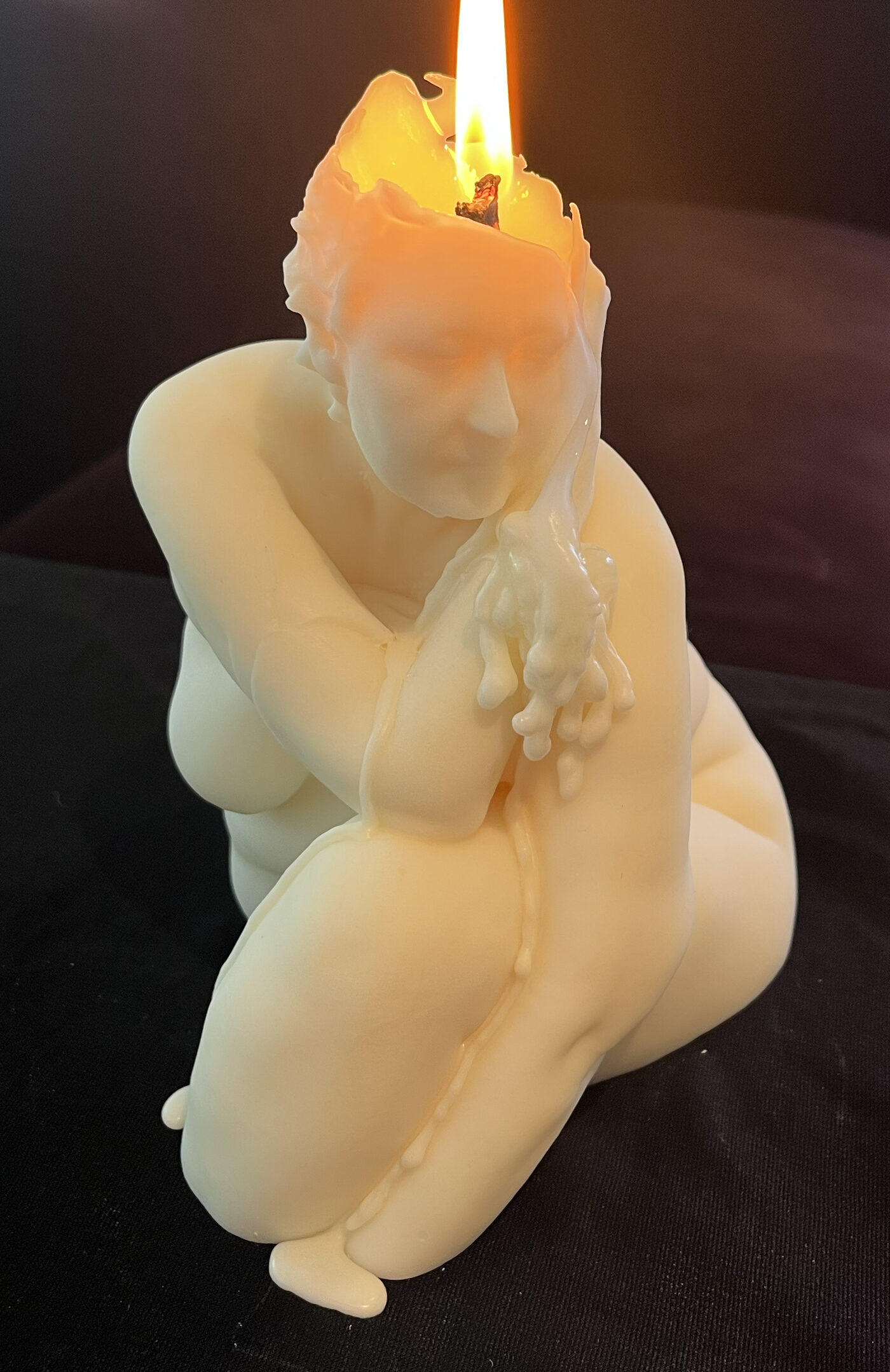 A pale wax sculpture of a seated figure, hunched forward, with a lit candle flame rising from the top of the head and wax dripping down the arms and hands.