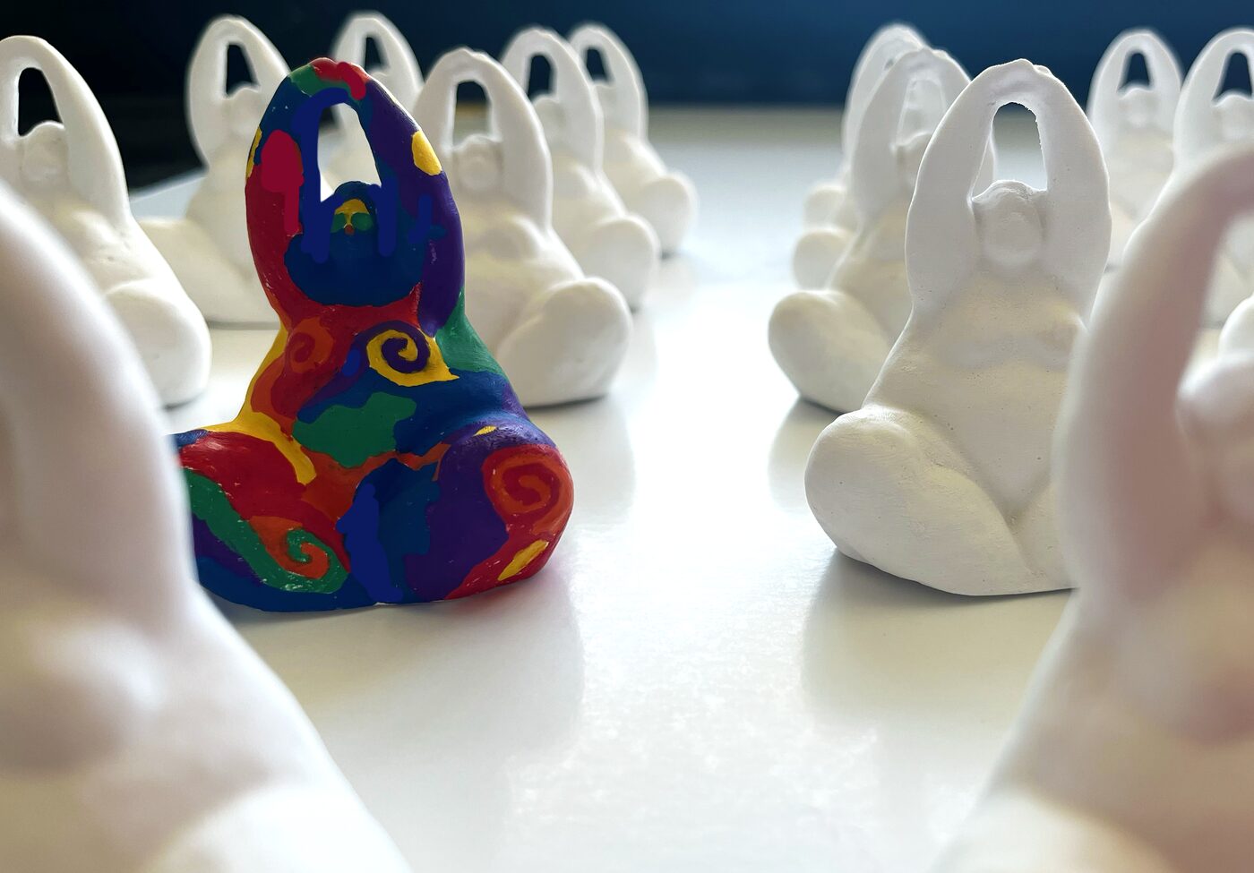 Rows of identical small white plaster figures, seen from behind, with one figure near the front hand-painted in bright, saturated colour.